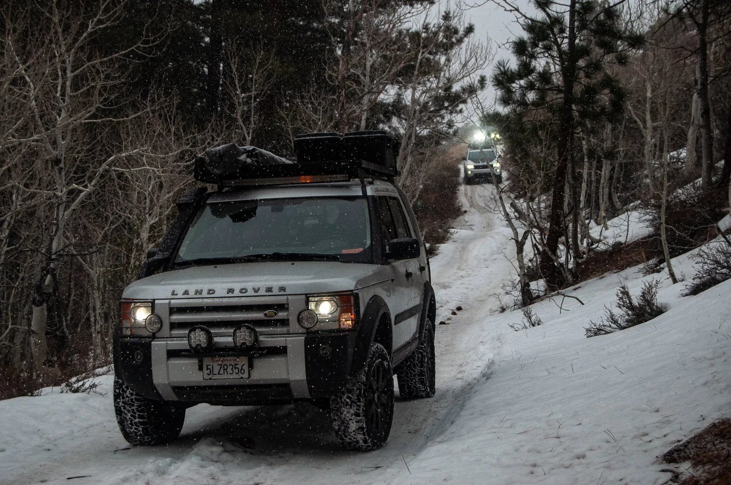 Two overland rigs driving a snowy forest trail