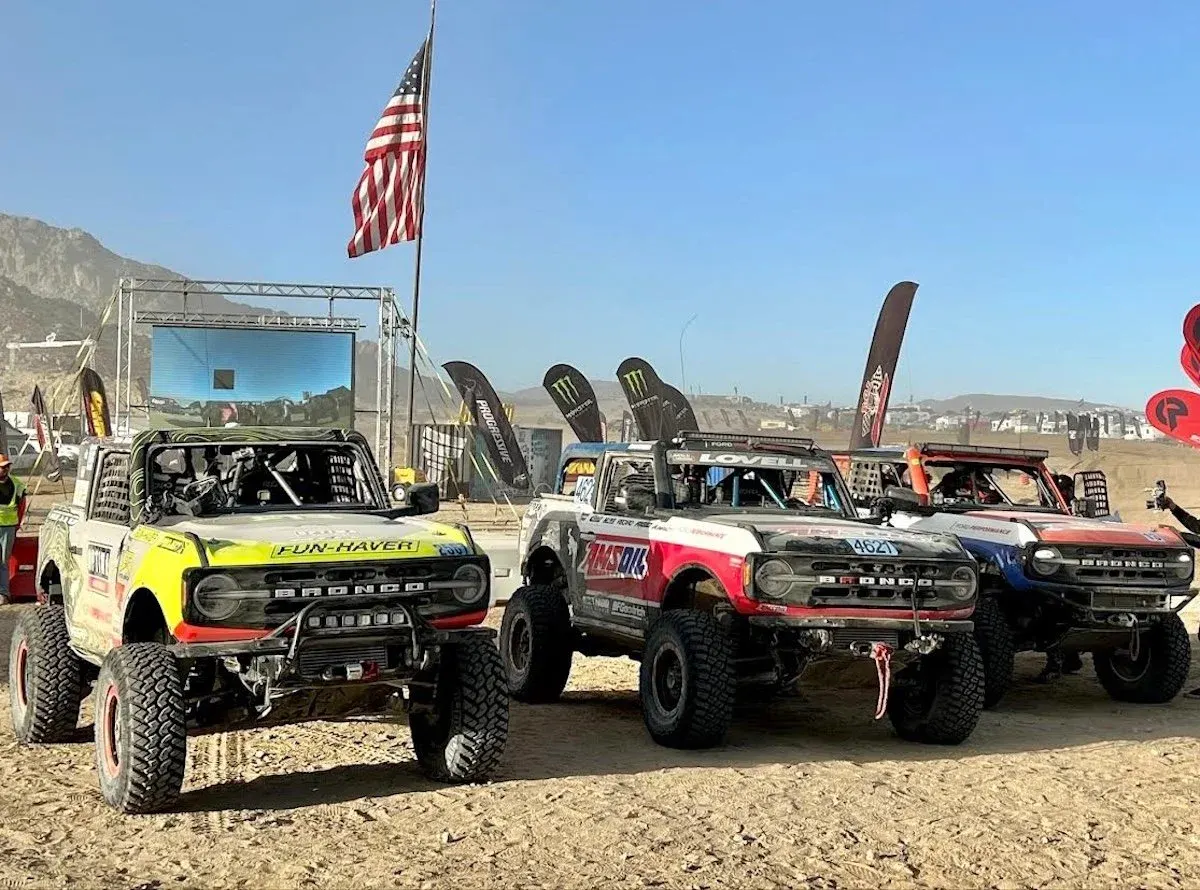 Ford Bronco race trucks lined up in the pits under an American flag
