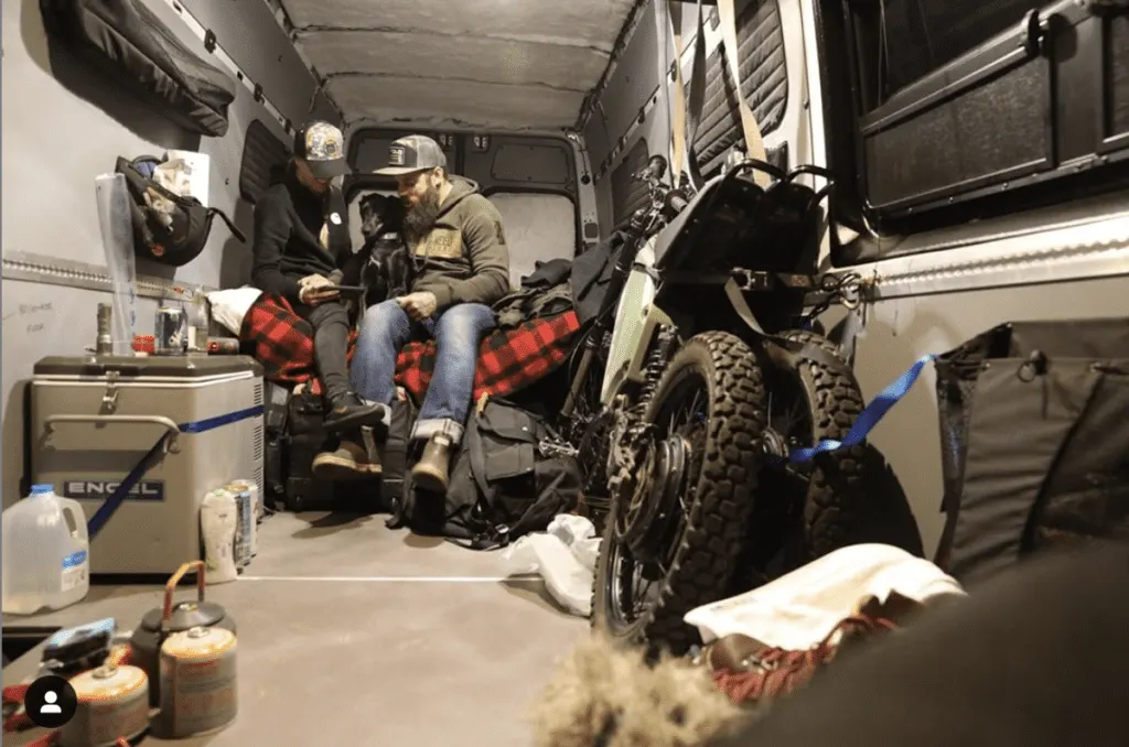 Two people sitting inside a cargo van with gear and a motorcycle