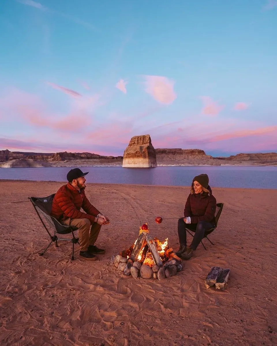 Two people sitting by a campfire on a lakeside beach at dusk
